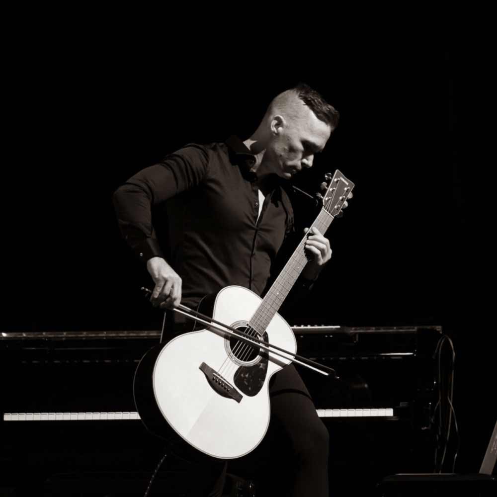 Black and white photograph of Timothy Fairless performing live, using a violin bow on an acoustic guitar.